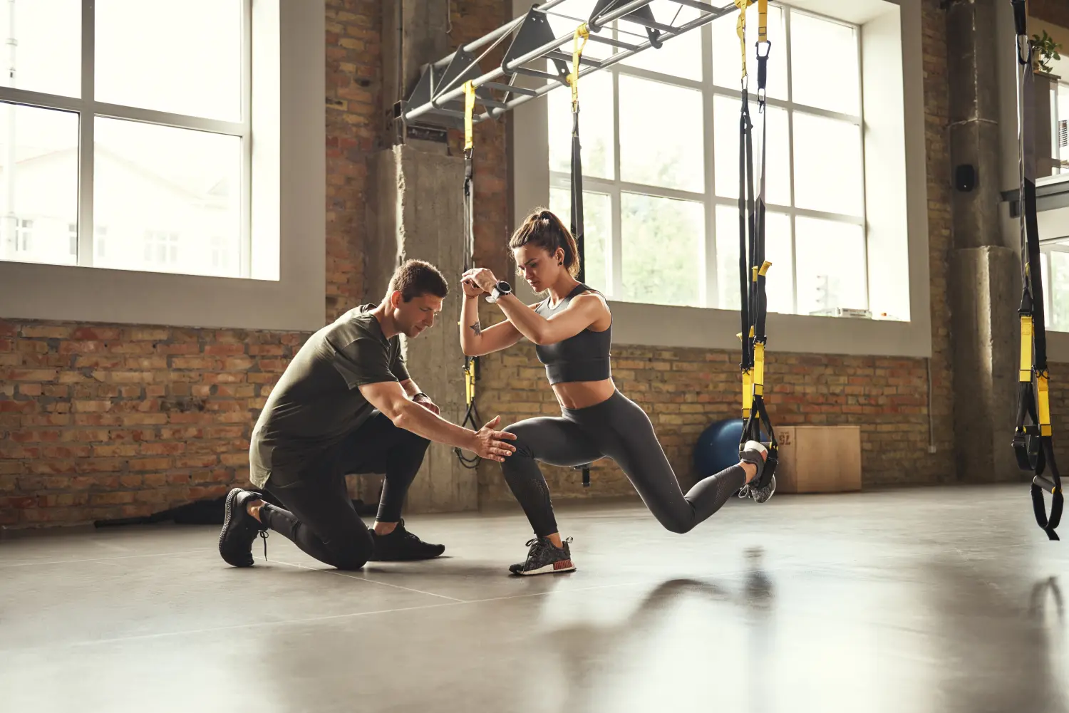 Selbstbewusster junger Personaltrainer zeigt einer schlanken, athletischen Frau, wie man beim Training im Fitnessstudio Kniebeugen mit Trx-Fitnessgurten macht.